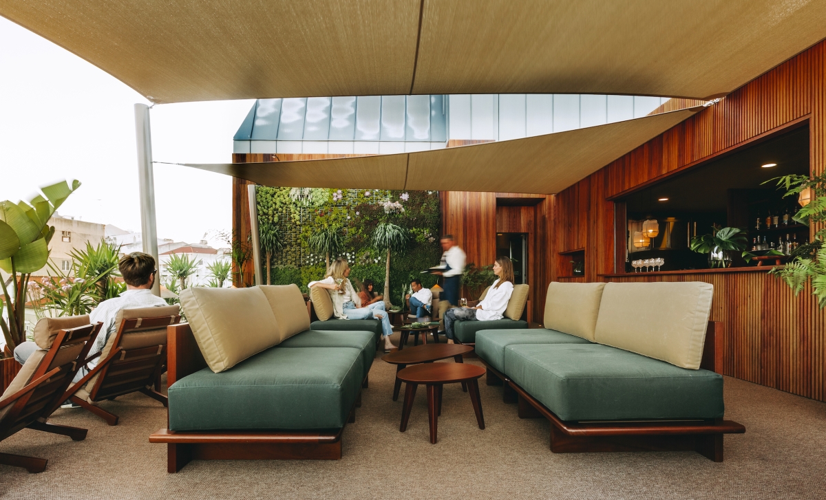 Des personnes se détendent sur des canapés d'extérieur modernes sous des parasols dans un salon de terrasse élégant avec des murs en bois, des plantes en pot et un bar à l'arrière-plan. Un serveur s'approche d'un groupe assis à une table.