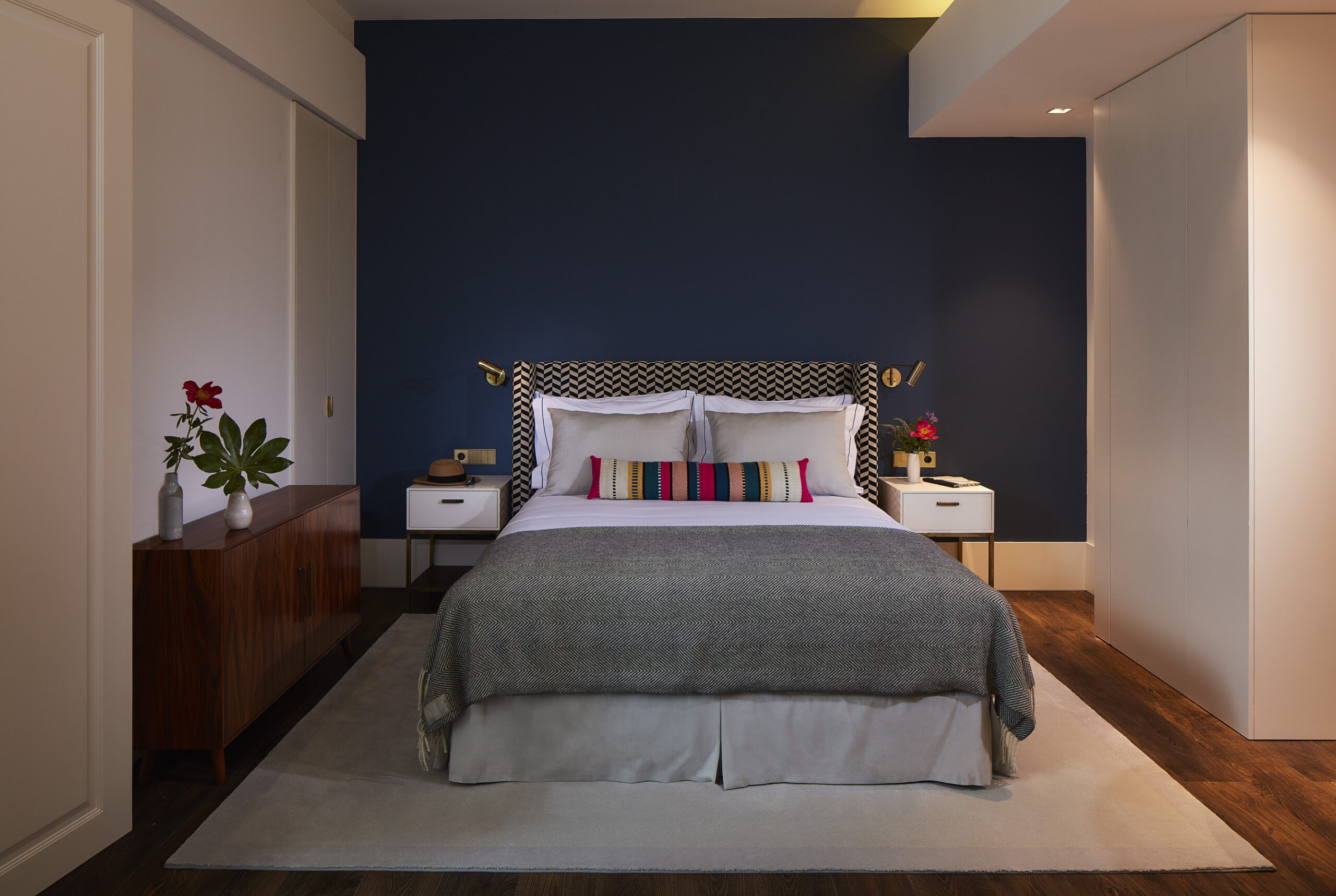 Un dormitorio moderno con una pared de acento azul, una cama con ropa de cama blanca y gris, almohadas de colores, dos mesillas de noche con lámparas, una cómoda de madera con jarrones y flores, y suelos de madera con una alfombra clara.