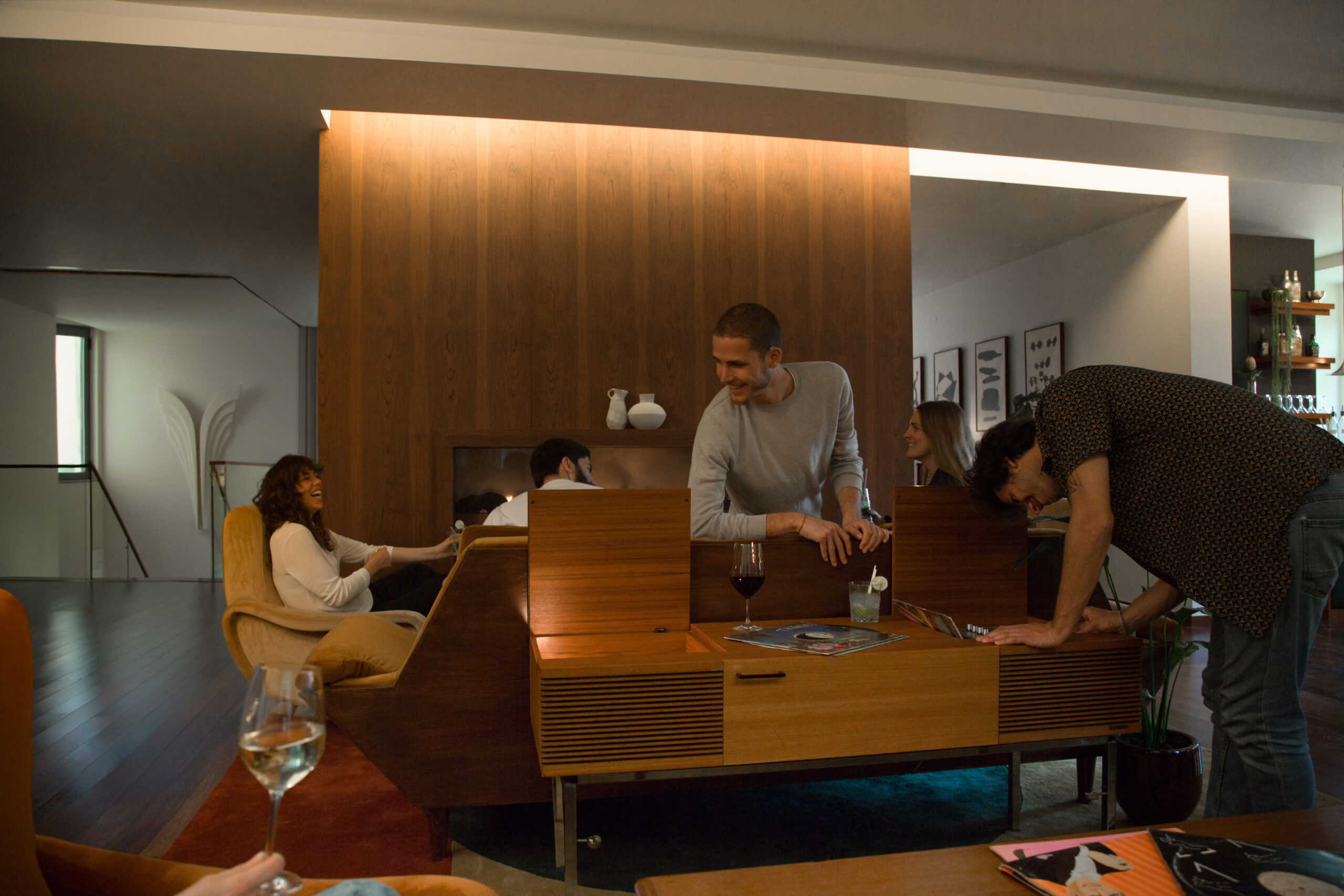 A group of friends relax and chat in a cozy, modern living room with wooden furniture; some are seated, while two men interact at a coffee table, with drinks and magazines visible.