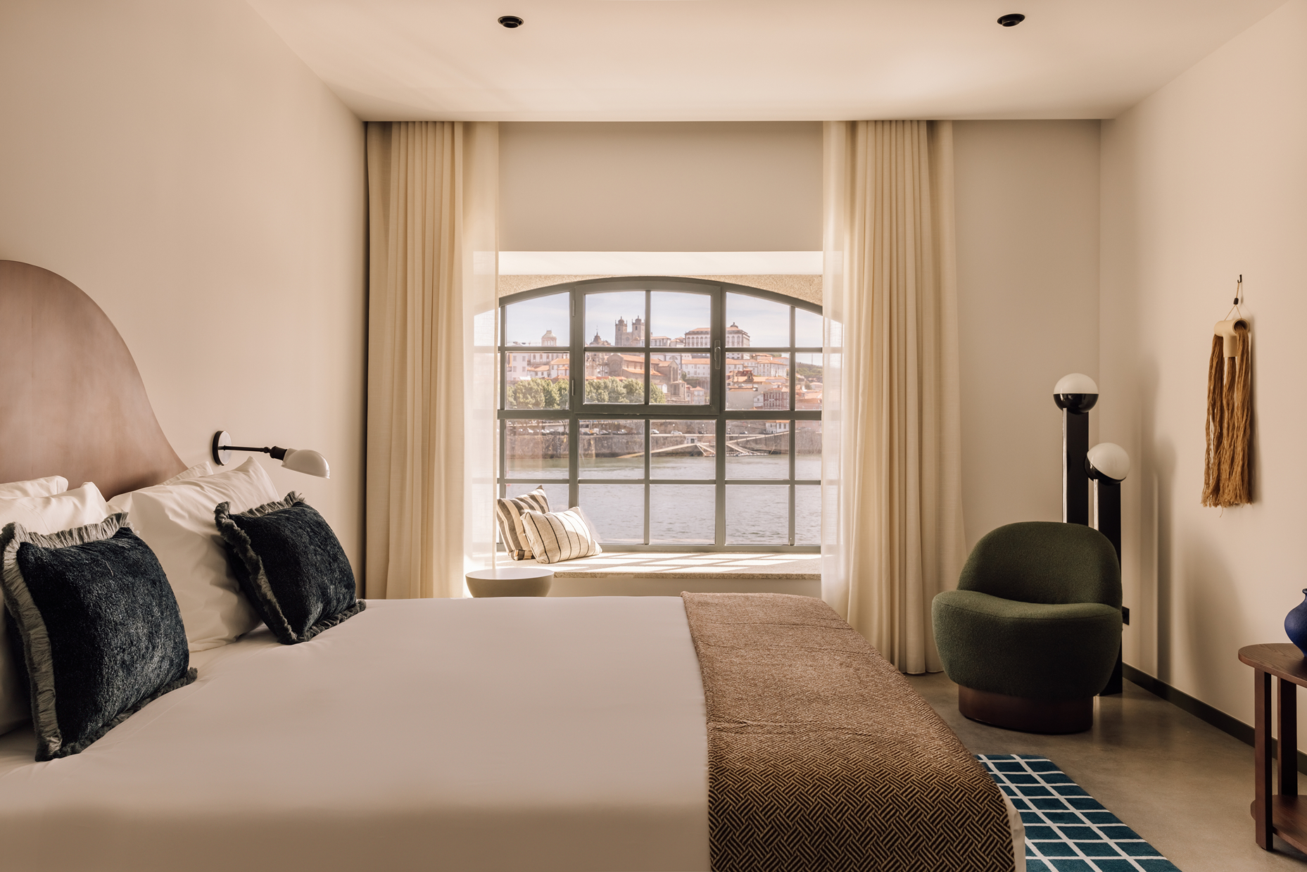 A modern hotel room with a large bed, dark blue cushions, green armchair, and a wide window offering a scenic river and city view. Neutral tones and natural light create a cozy, inviting atmosphere.