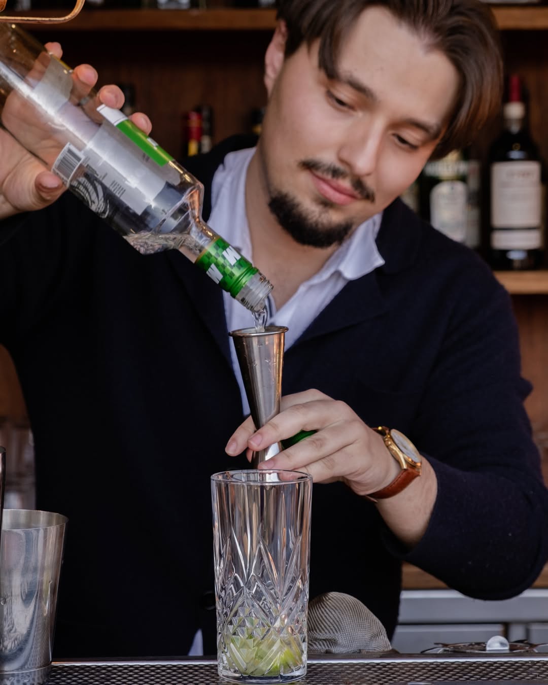 Un camarero con vello facial vierte líquido transparente de una botella en un jigger metálico sobre un vaso de cristal con gajos de lima, de pie en una barra con botellas al fondo.
