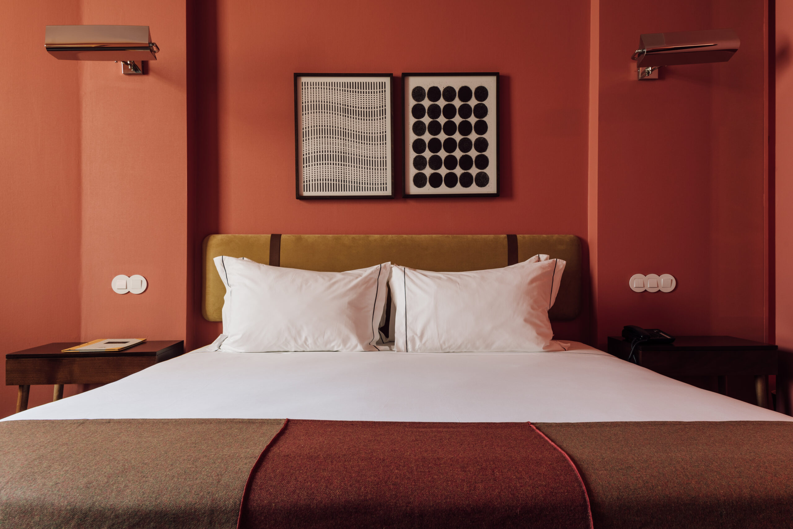 A neatly made bed with white pillows and sheets is set against a warm, red-toned wall, flanked by two side tables, with two abstract framed artworks hanging above the headboard.