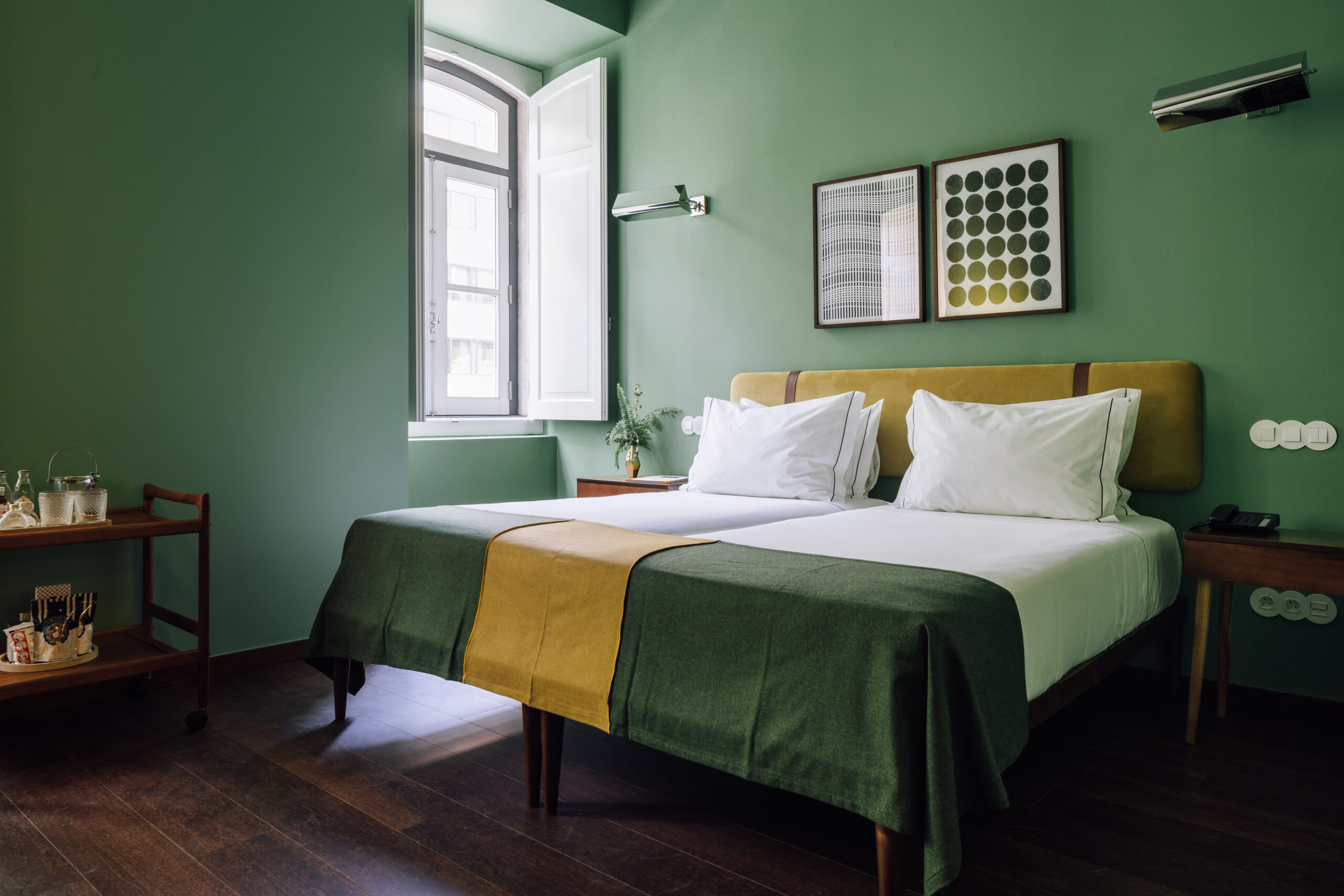 A modern hotel room with green walls, a double bed with white pillows and green and yellow bedding, two framed artworks above the headboard, a window letting in natural light, and a wooden cart beside the bed.