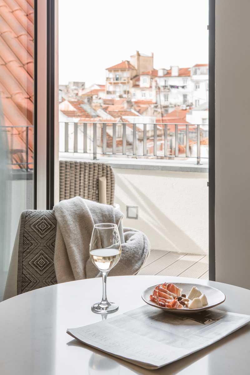 A glass of white wine and a plate with cheese and prosciutto sit on a table by a newspaper, with a cozy chair and a view of city rooftops through a balcony window in the background.