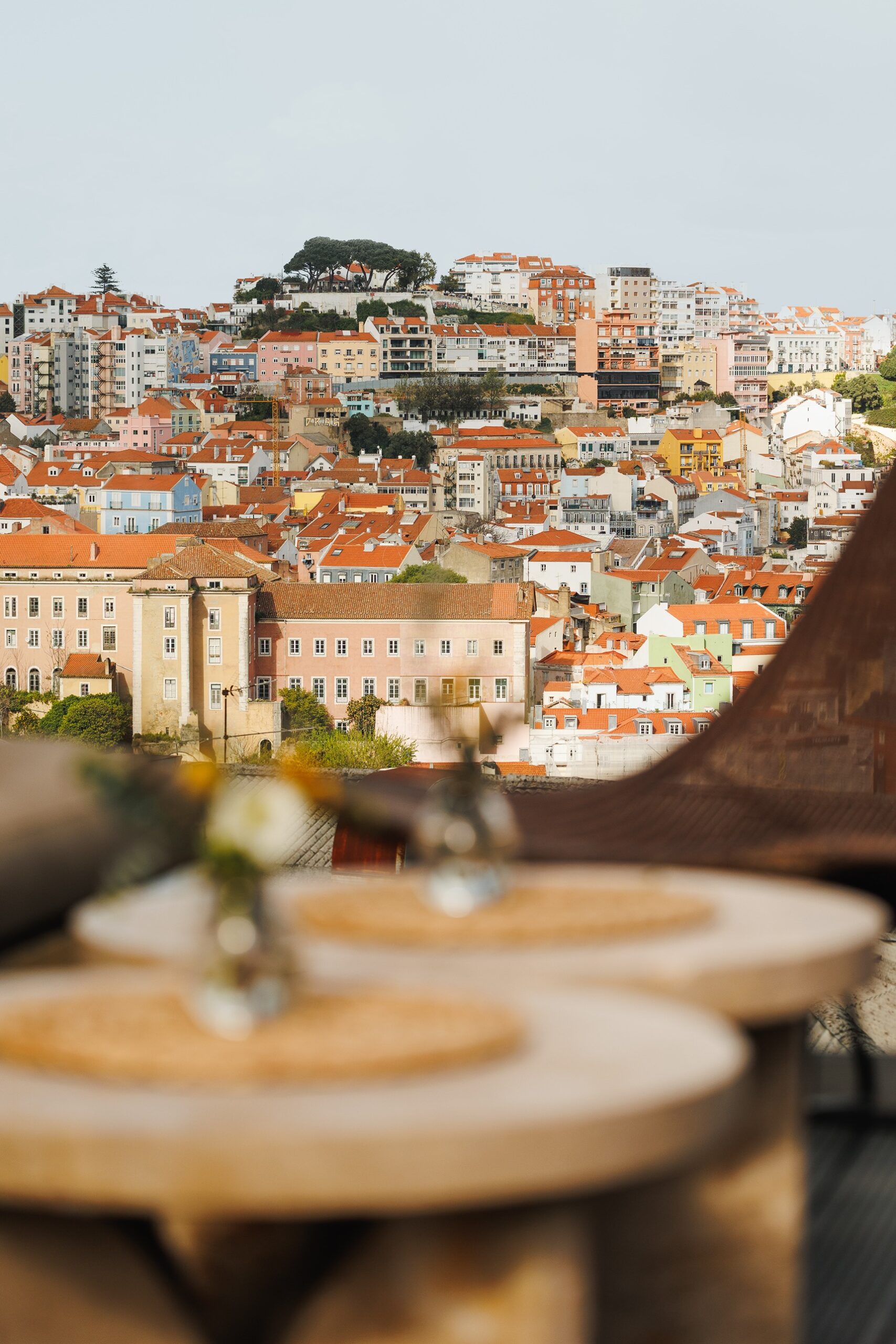 A scenic view of a colorful hillside city, likely Lisbon, featuring clusters of pastel buildings with red roofs; blurred table settings are visible in the foreground.
