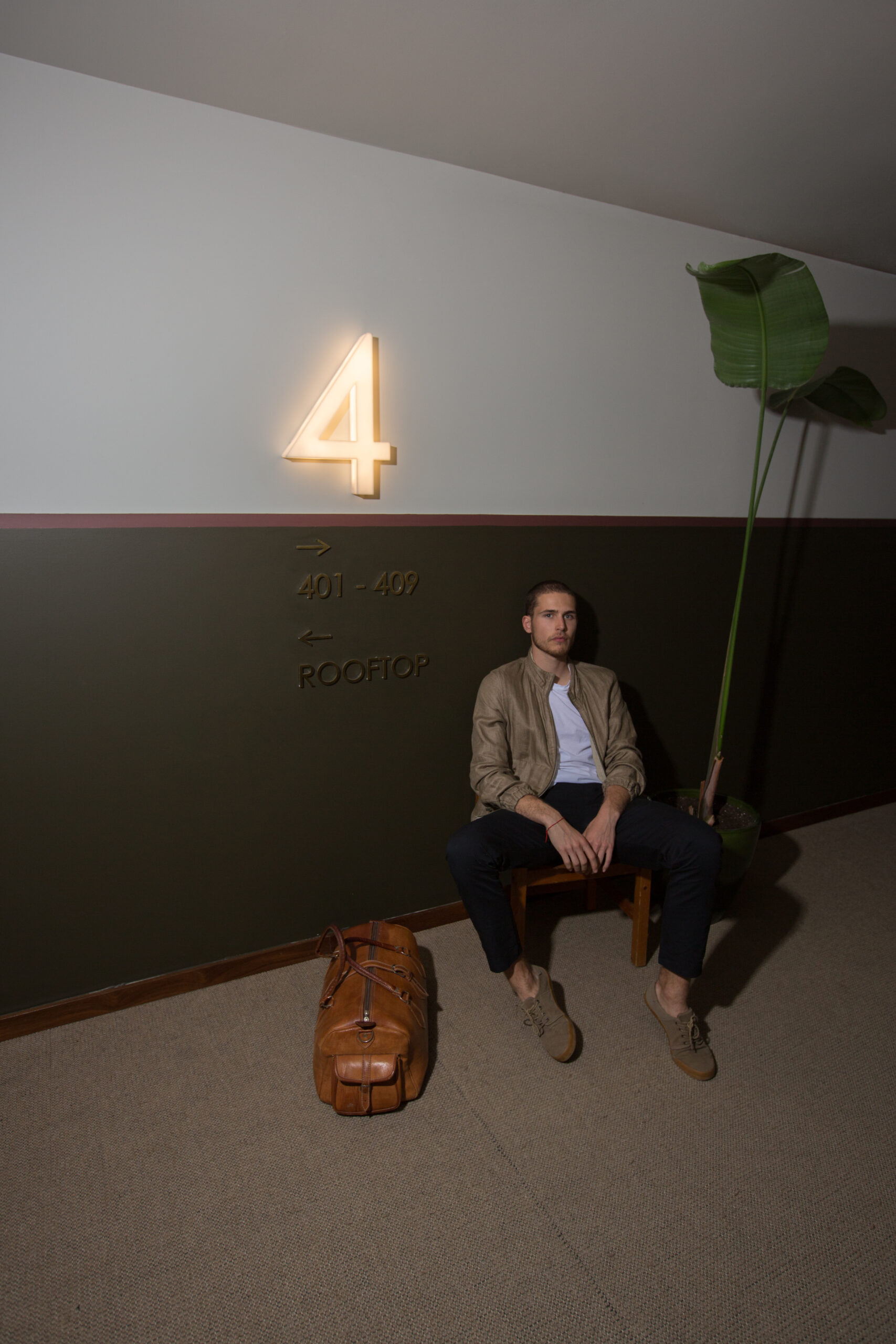 Un homme est assis sur un banc à côté d'une plante en pot et d'un grand sac de sport marron sous un panneau lumineux indiquant le numéro 4, avec un texte mural indiquant les directions vers les chambres 401-409 et un toit.