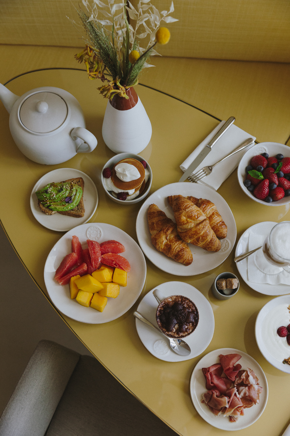Un petit-déjeuner sur une table jaune comprenant des croissants, des tranches de mangue et de pamplemousse, des toasts à l'avocat, des baies, de la confiture, du prosciutto, une théière, une tasse de café, un yaourt avec des fruits et un vase avec des fleurs séchées.