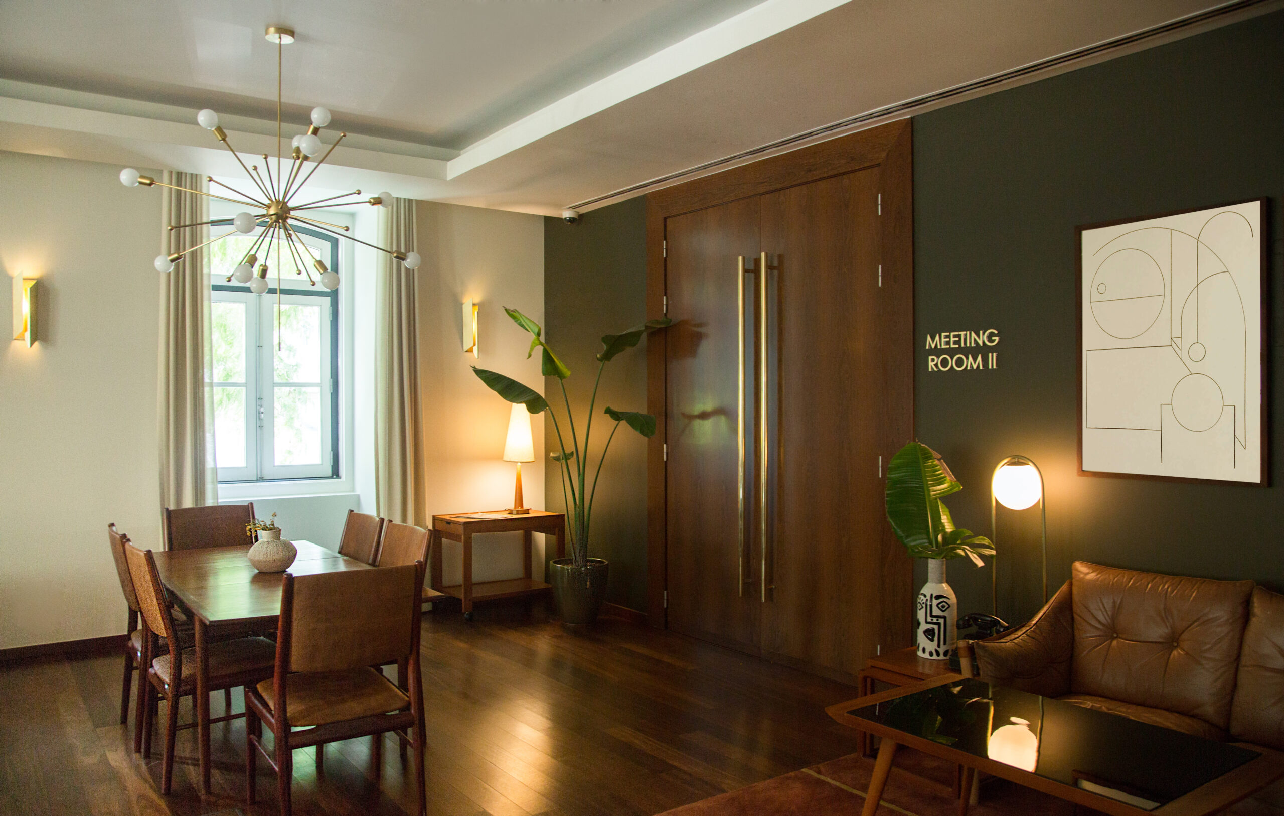 A modern meeting room with a wooden table and six chairs, green walls, large wooden doors, indoor plants, wall art, and soft lighting from lamps and a ceiling fixture. 