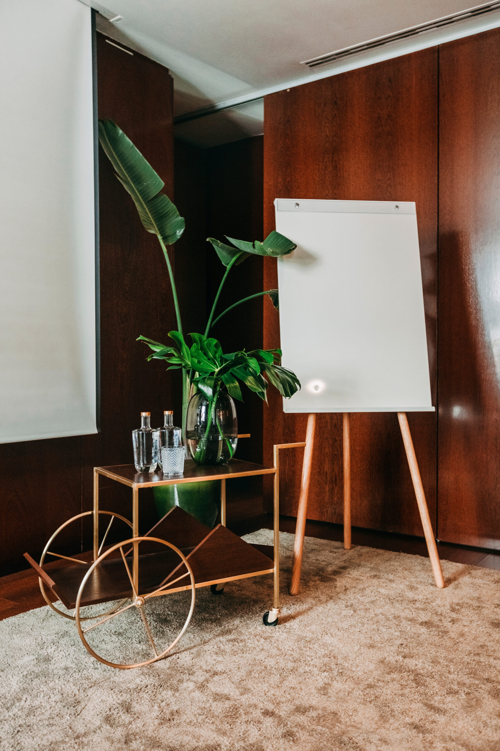 A blank flip chart stands on an easel next to a gold cart holding bottled water and a large green plant in a modern, wood-paneled room with beige carpet.