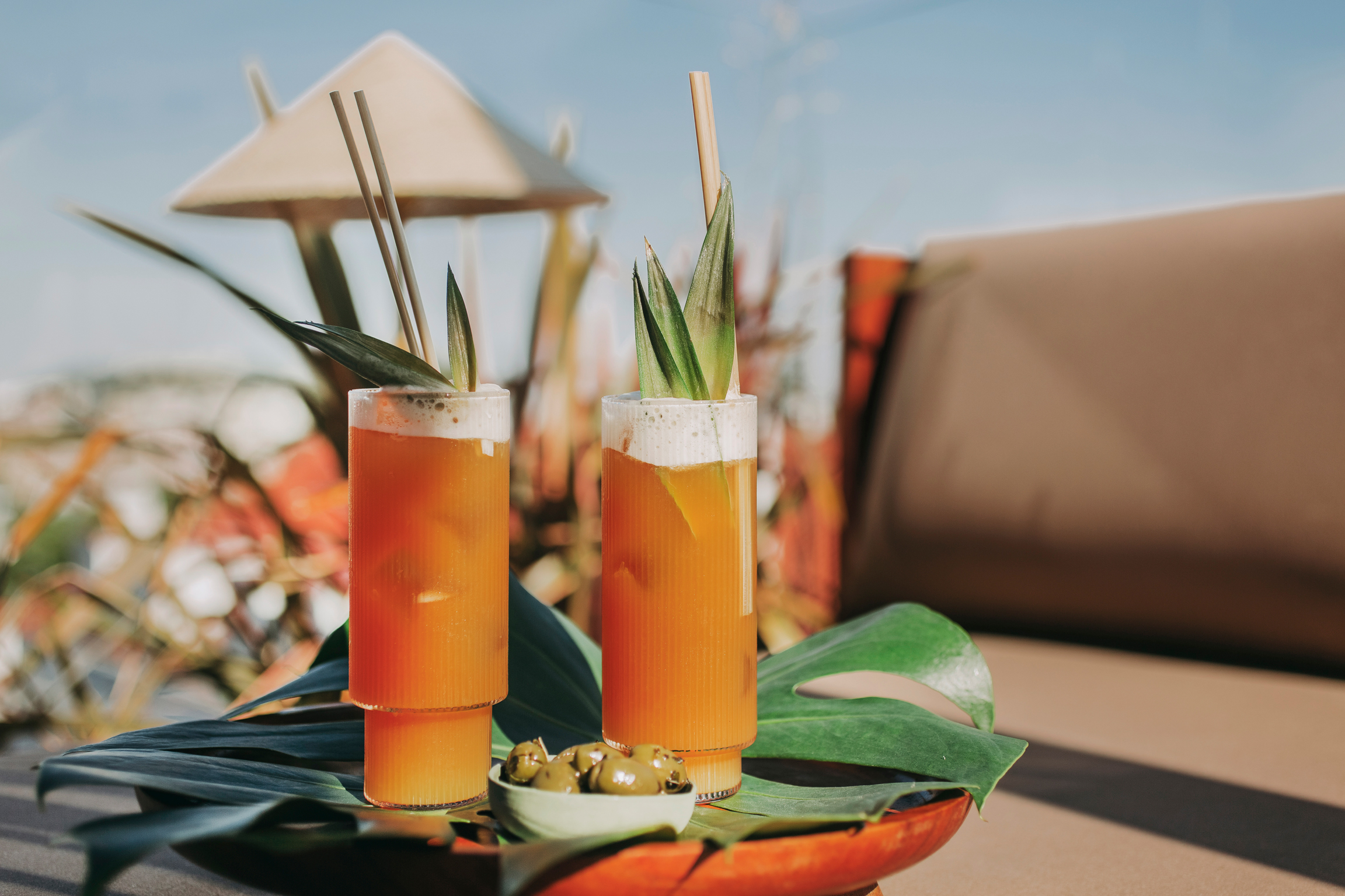 Deux grands verres de cocktails tropicaux orange avec garniture ananas et pailles sont posés sur des feuilles vertes, à côté d'un petit bol d'olives, en extérieur avec un arrière-plan ensoleillé flou et un parasol en paille.