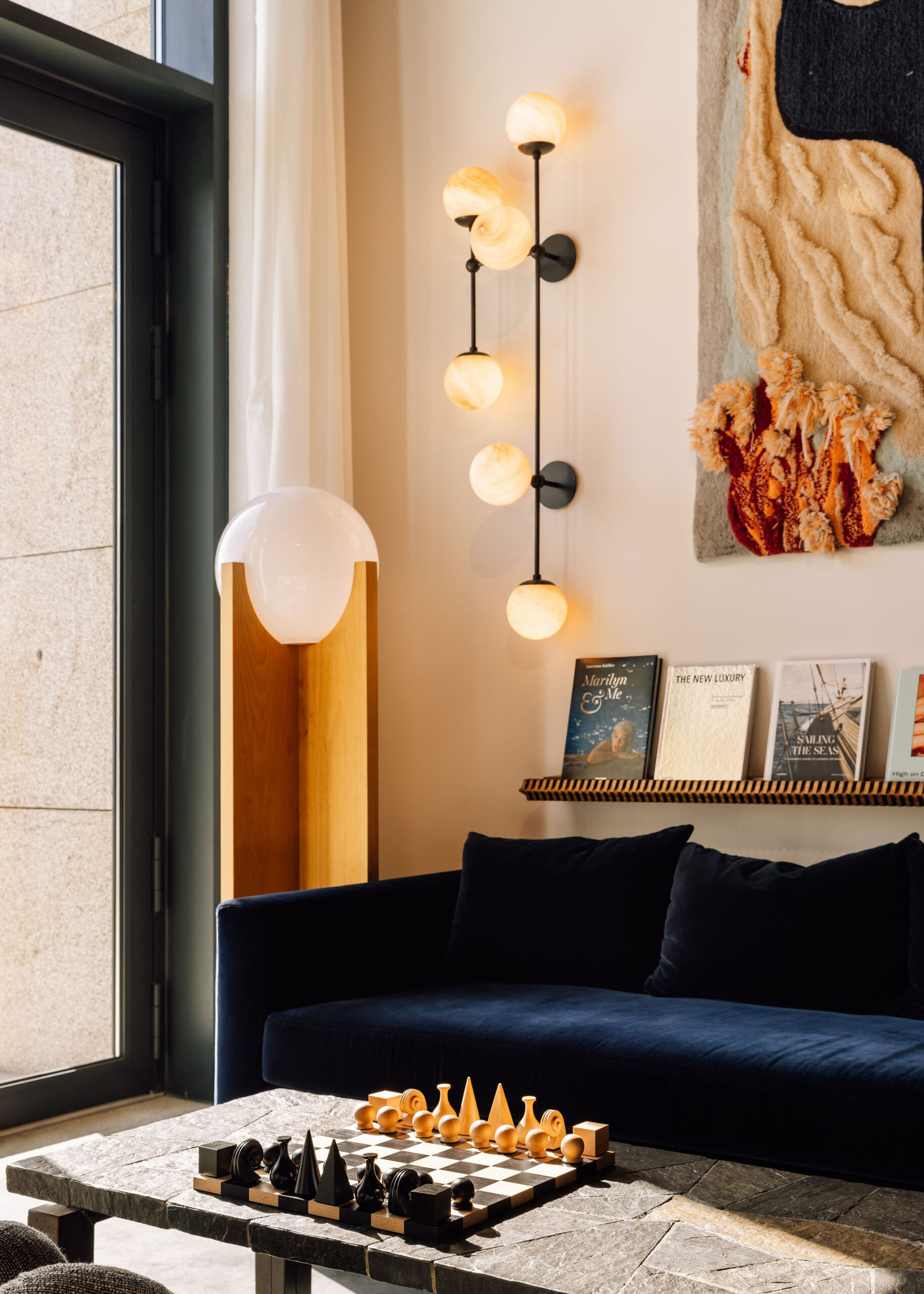 A cozy modern living room with a navy sofa, a unique vertical wall lamp with round bulbs, a stone coffee table with a sculptural chess set, a wooden lamp, a magazine rack, and textile wall art. Sunlight streams through a large window.
