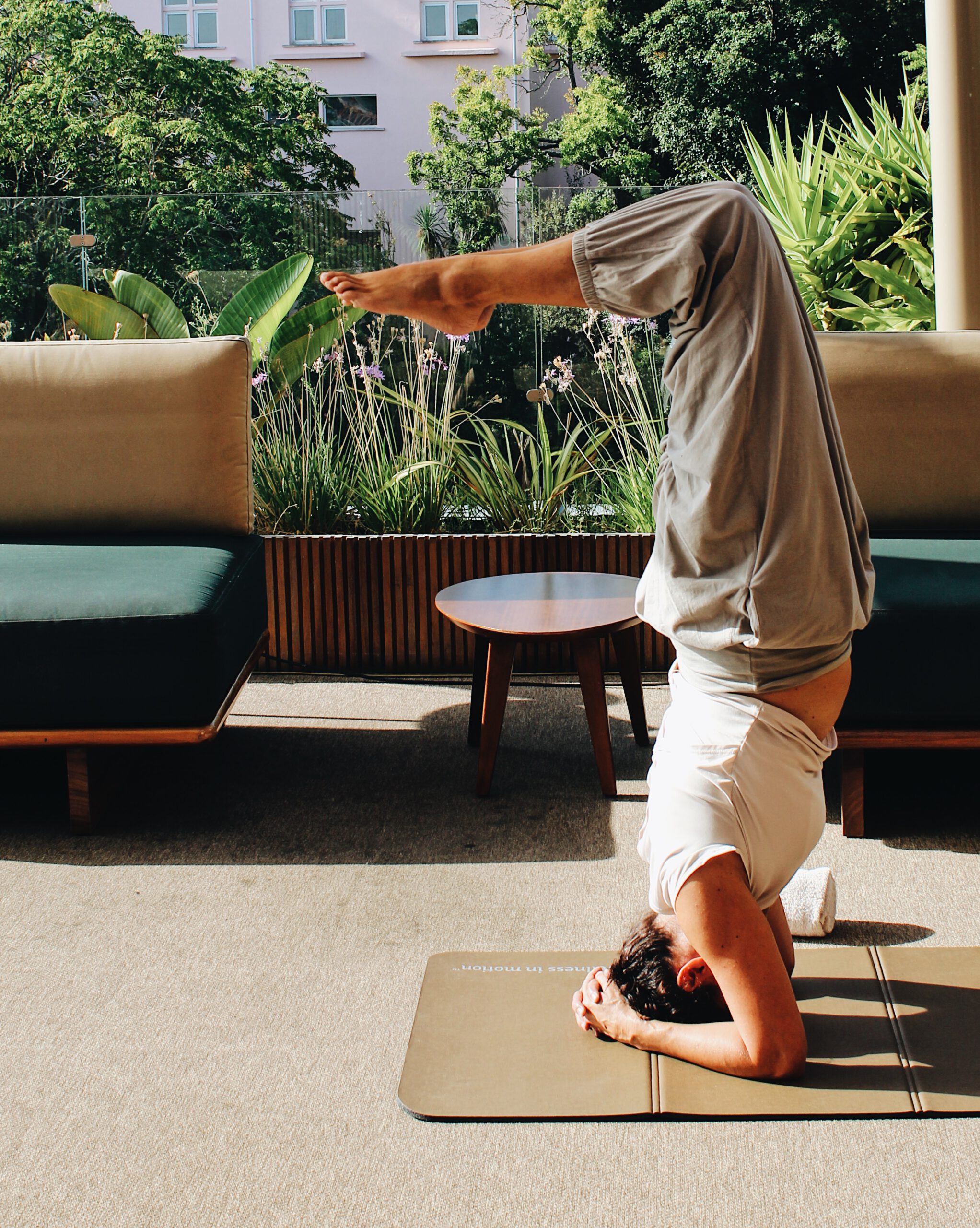 A person practices a yoga headstand on a mat indoors, with legs bent and toes pointed up. Sunlight streams in, and greenery and sofas are visible in the background.