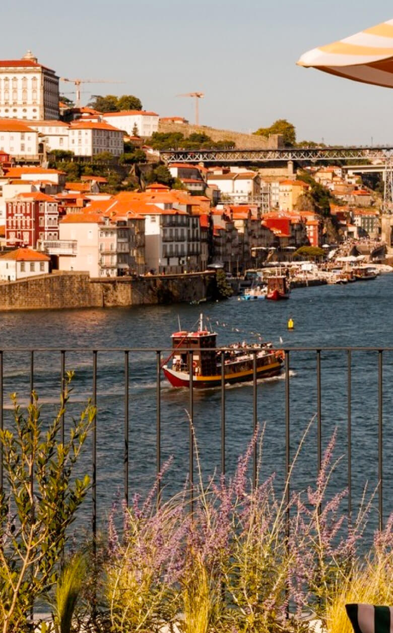 A scenic riverside view of Porto, Portugal, showing colorful buildings, a traditional boat on the Douro River, and a bridge in the background, with plants and a railing in the foreground.