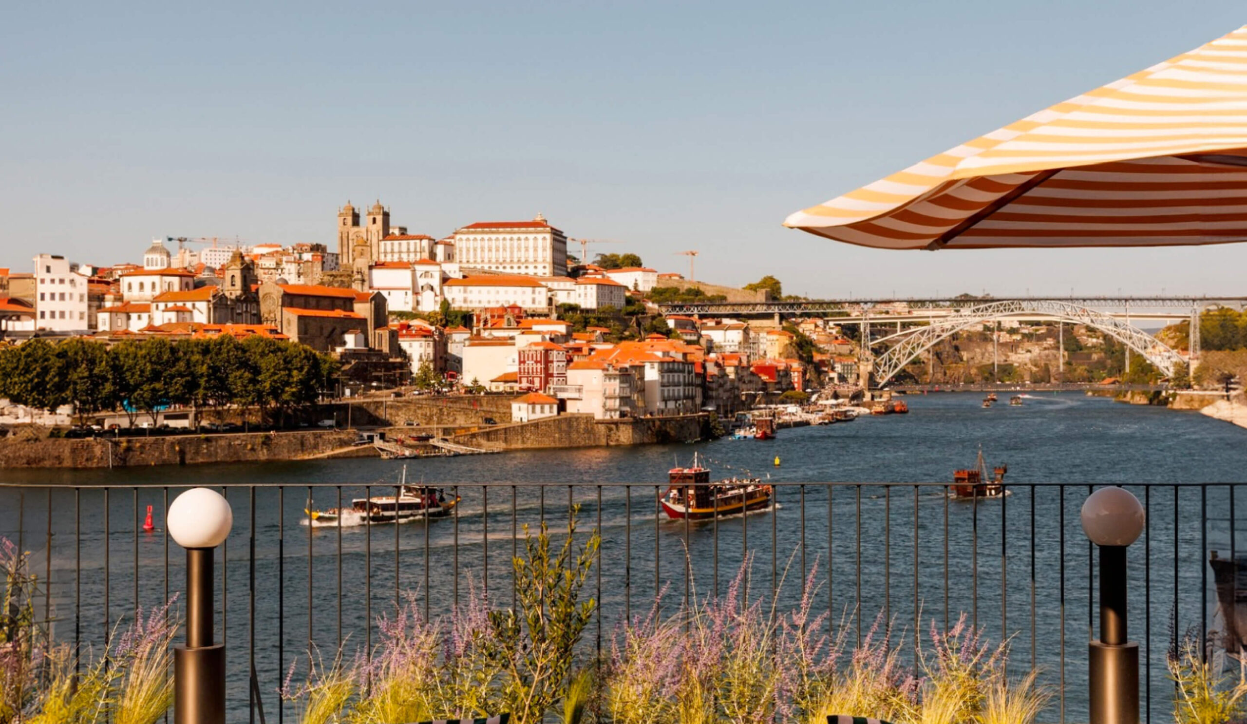 View of a river with several boats, a metal bridge in the background, and a hillside city with red-roofed buildings, seen from a terrace under a striped umbrella on a sunny day.