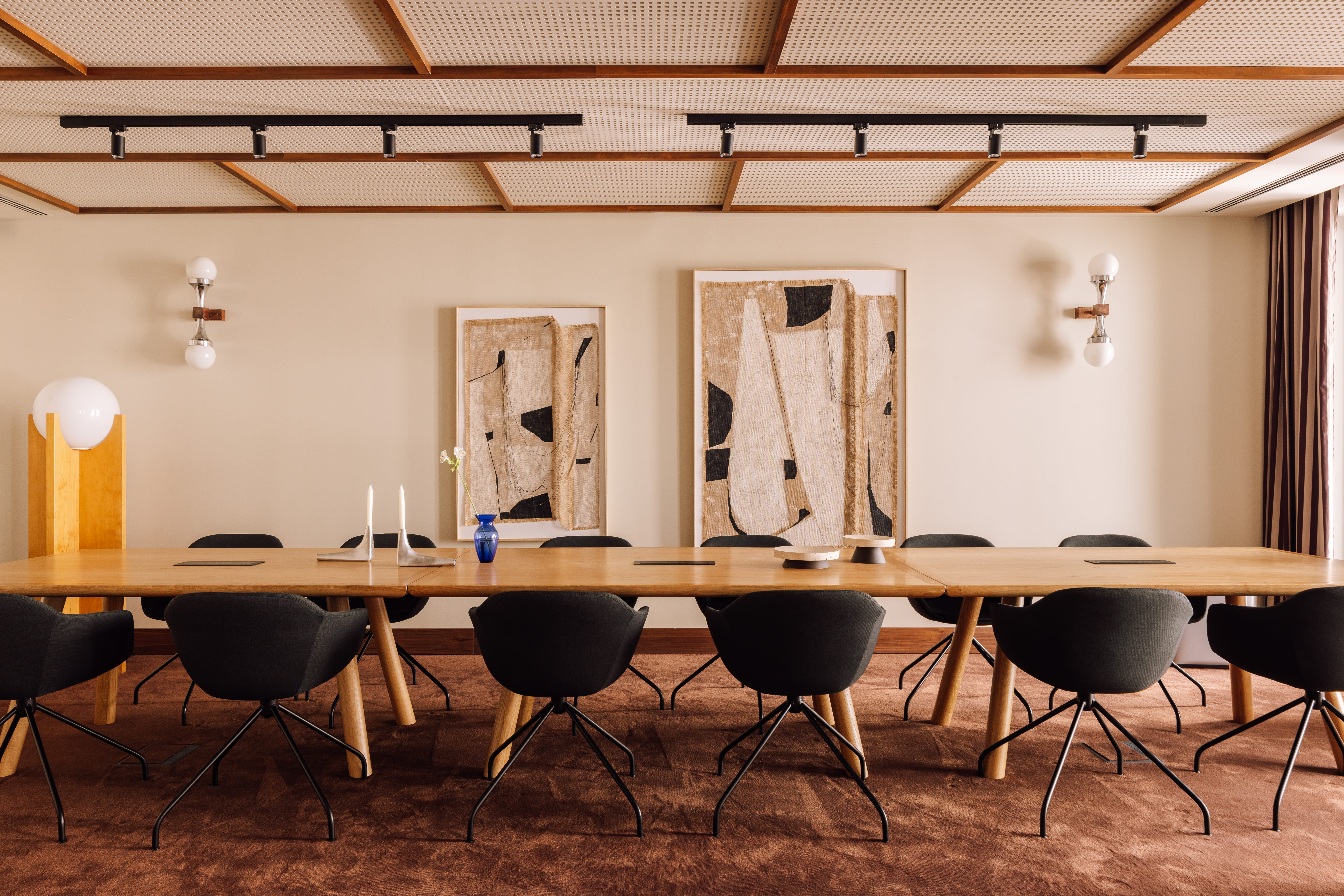 A modern conference room with a long wooden table, black chairs, abstract art on the wall, soft lighting from wall sconces, and a brown carpeted floor. Minimalist decor includes candles and a vase on the table.
