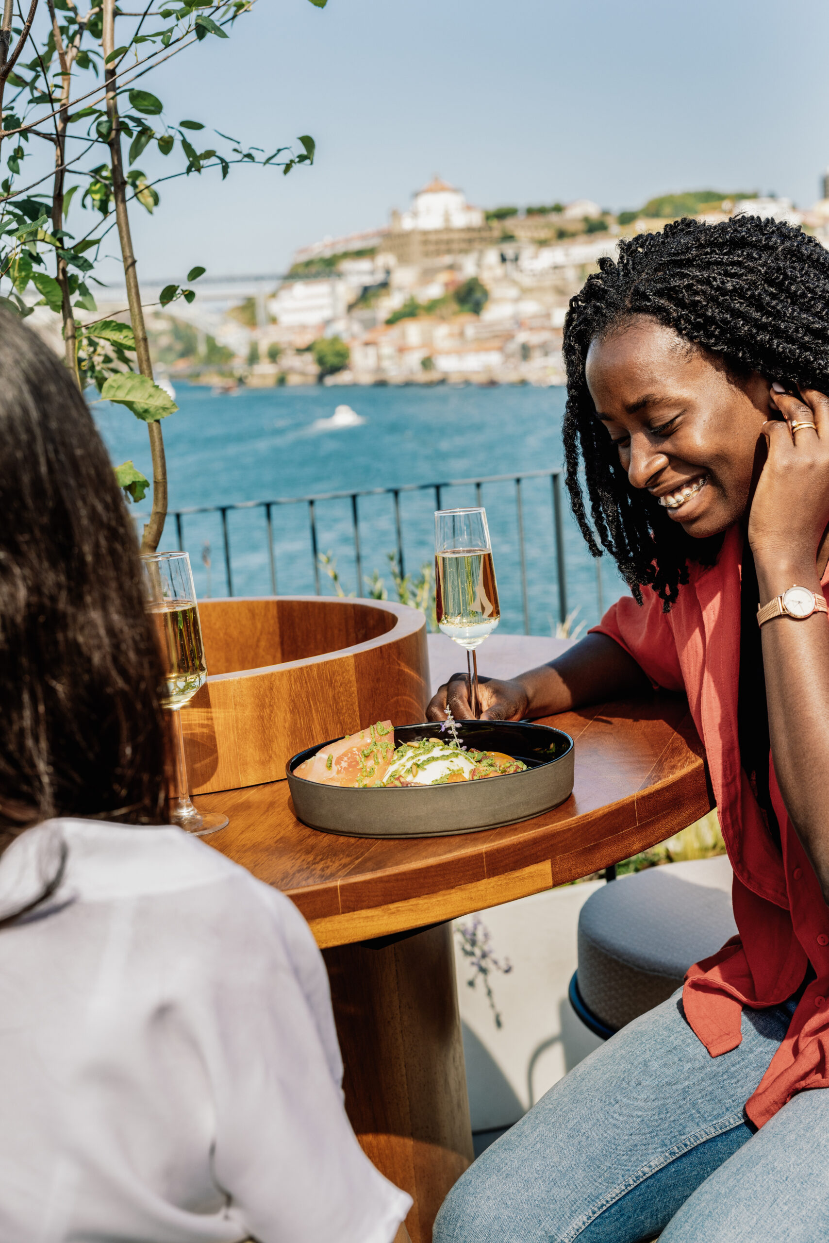 Two women sit at an outdoor table by the water, enjoying drinks and a meal. One woman, smiling, wears a red shirt, while the other faces away. Sunny weather and scenic buildings are visible in the background.