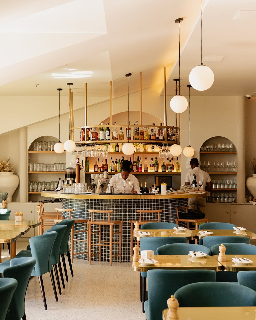 Un restaurant moderne et lumineux avec des chaises vertes, des tables en bois et un bar central. Deux personnes travaillent derrière le bar, qui est garni de bouteilles et de verres. Des suspensions rondes sont accrochées au plafond.