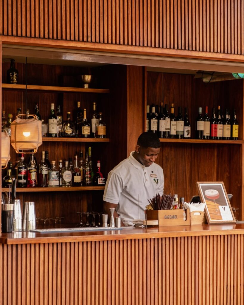 Un barman en chemise blanche se tient derrière un bar en bois, entouré d'étagères de bouteilles et de verres, et prépare des boissons dans un cadre moderne et chaleureusement éclairé.