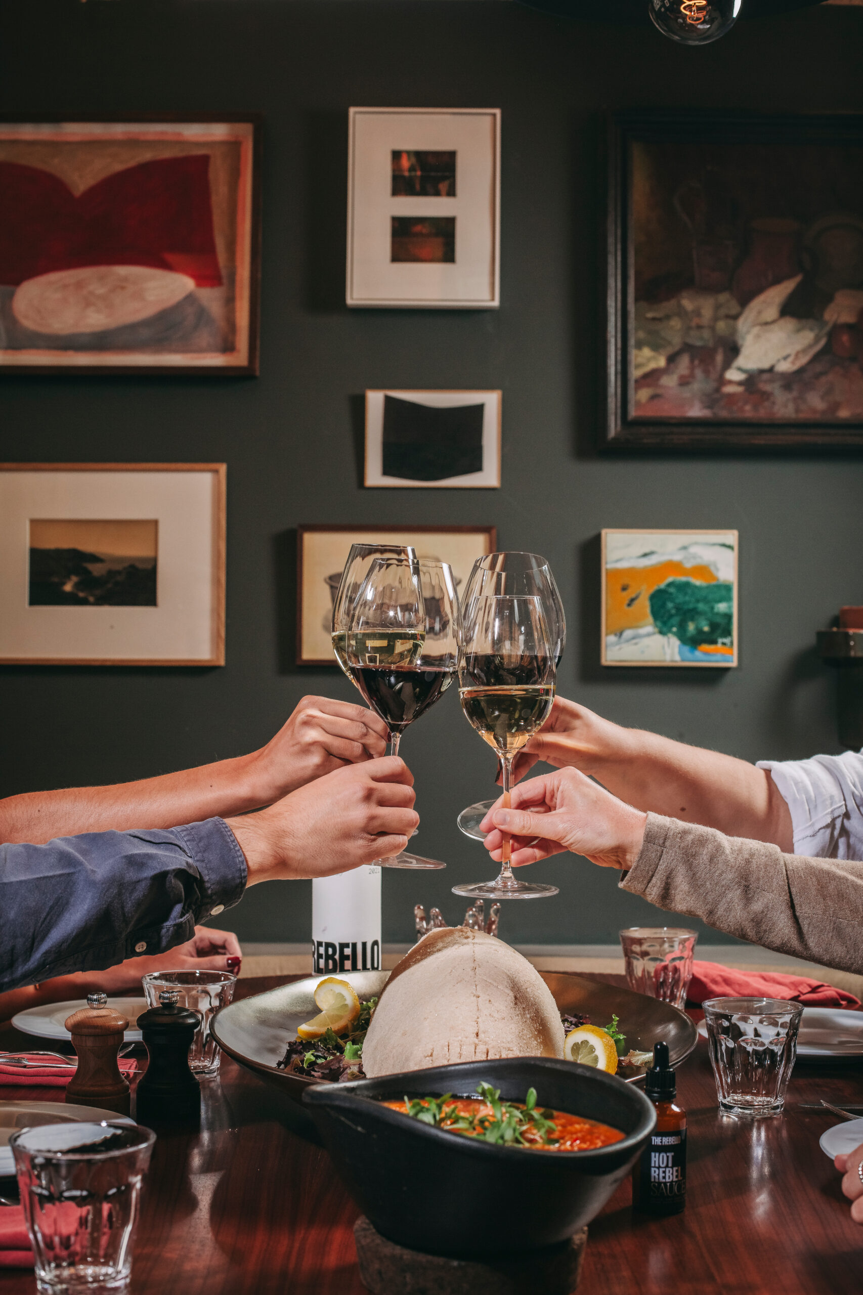 Four people clink wine glasses over a dinner table with food and drinks; the background features a wall decorated with framed artwork.