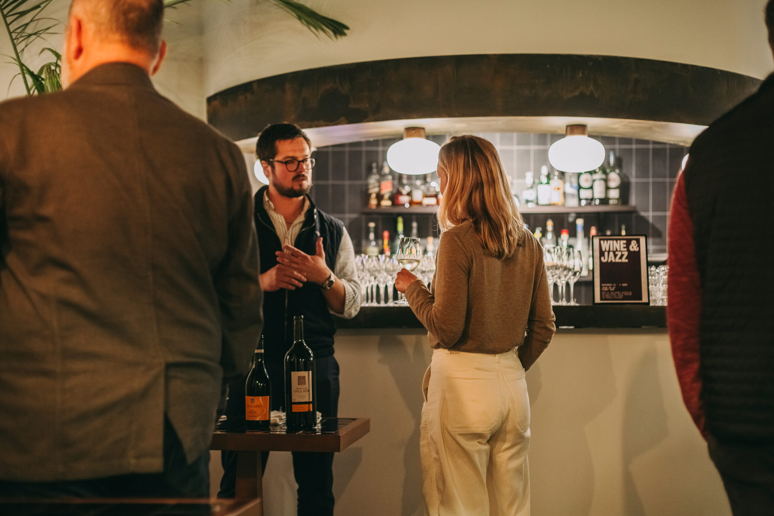 A man and woman stand and converse near a bar lined with glasses and bottles. Other people are in the foreground. A “Wine & Jazz” sign is visible on the counter. The setting appears warm and social.