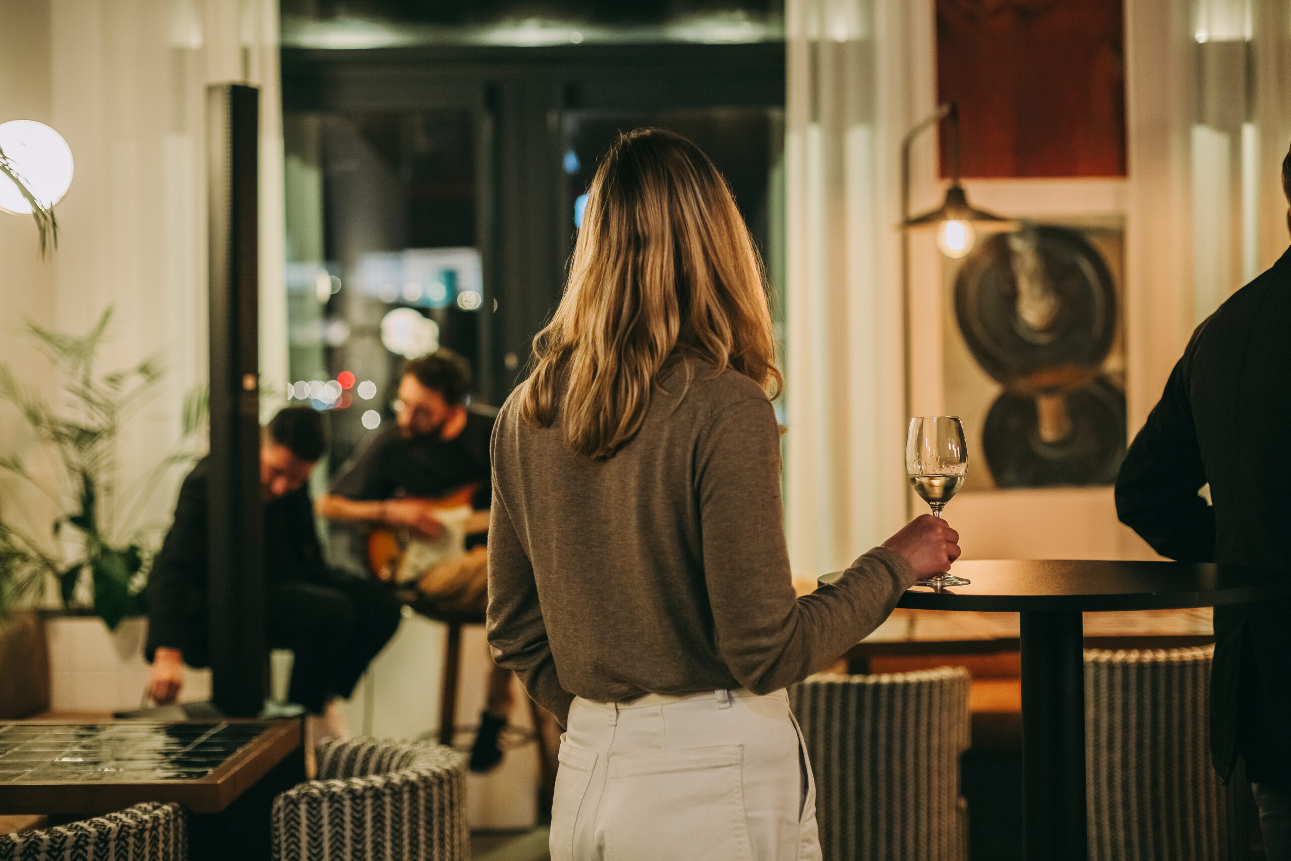 A woman with blonde hair, seen from behind, stands holding a glass of wine in a cozy, dimly lit lounge, while two musicians perform in the background near a window.