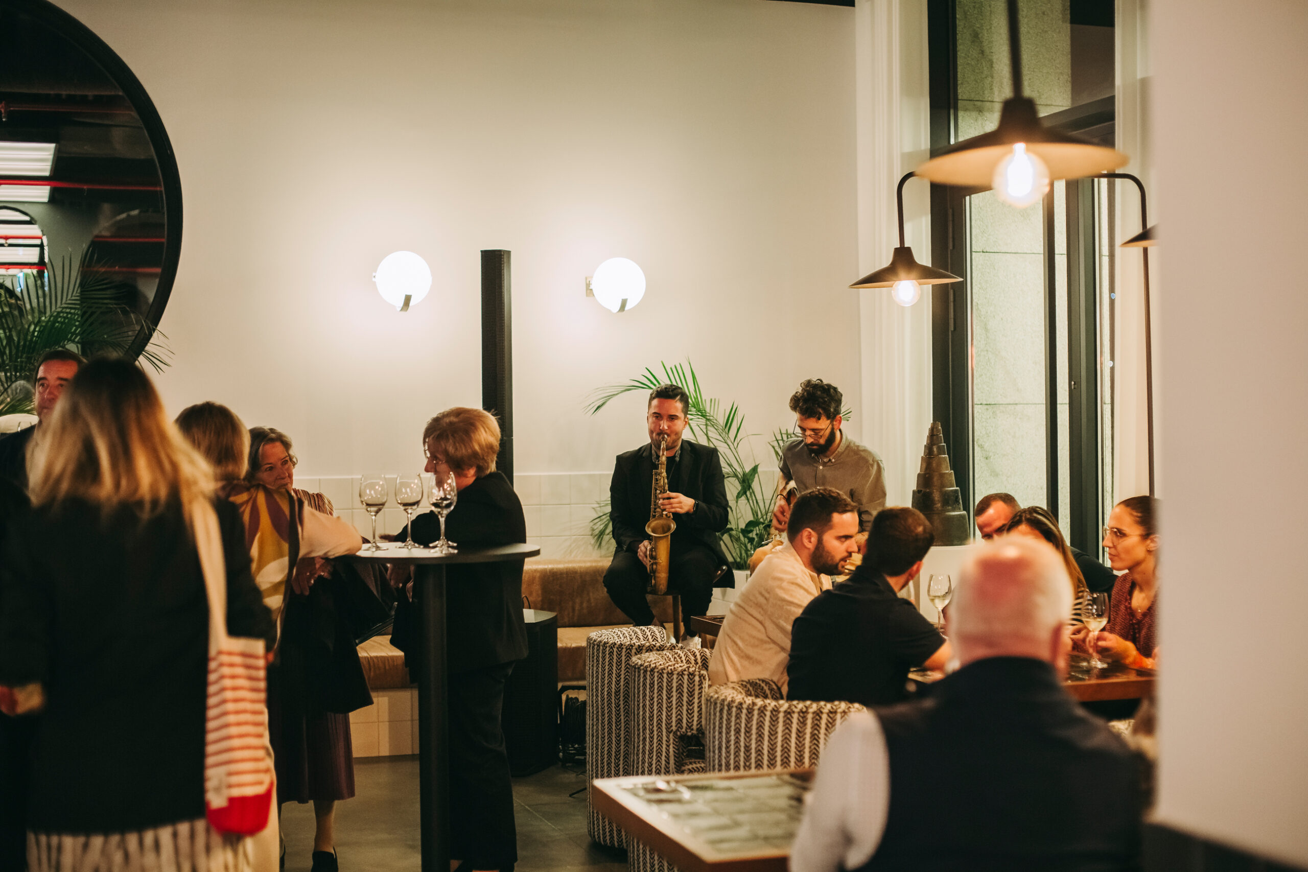 People gather in a stylish restaurant with modern lighting. A server carries drinks, and a musician plays a saxophone near the back. Some guests are seated and chatting while others stand and mingle.