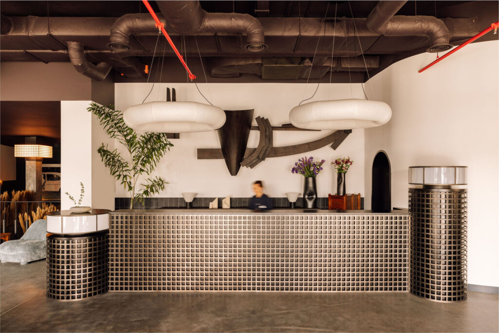 A modern hotel reception desk with a geometric tile front, two large round white hanging lights, sculptures and plants behind the desk, and a blurred figure standing at the counter.