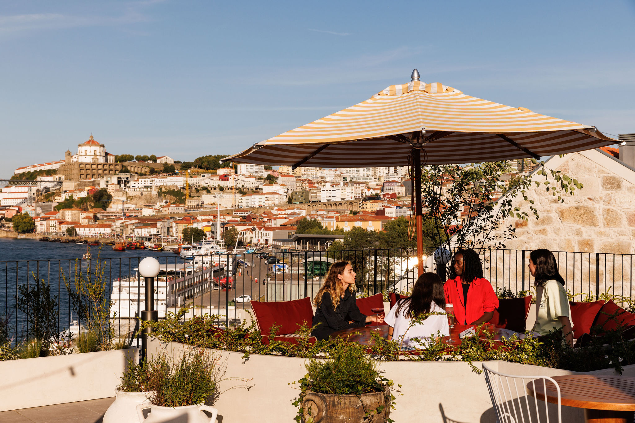 Four women sit talking at an outdoor terrace under a striped umbrella, overlooking a river and a hillside city with historic buildings in the background on a sunny day.