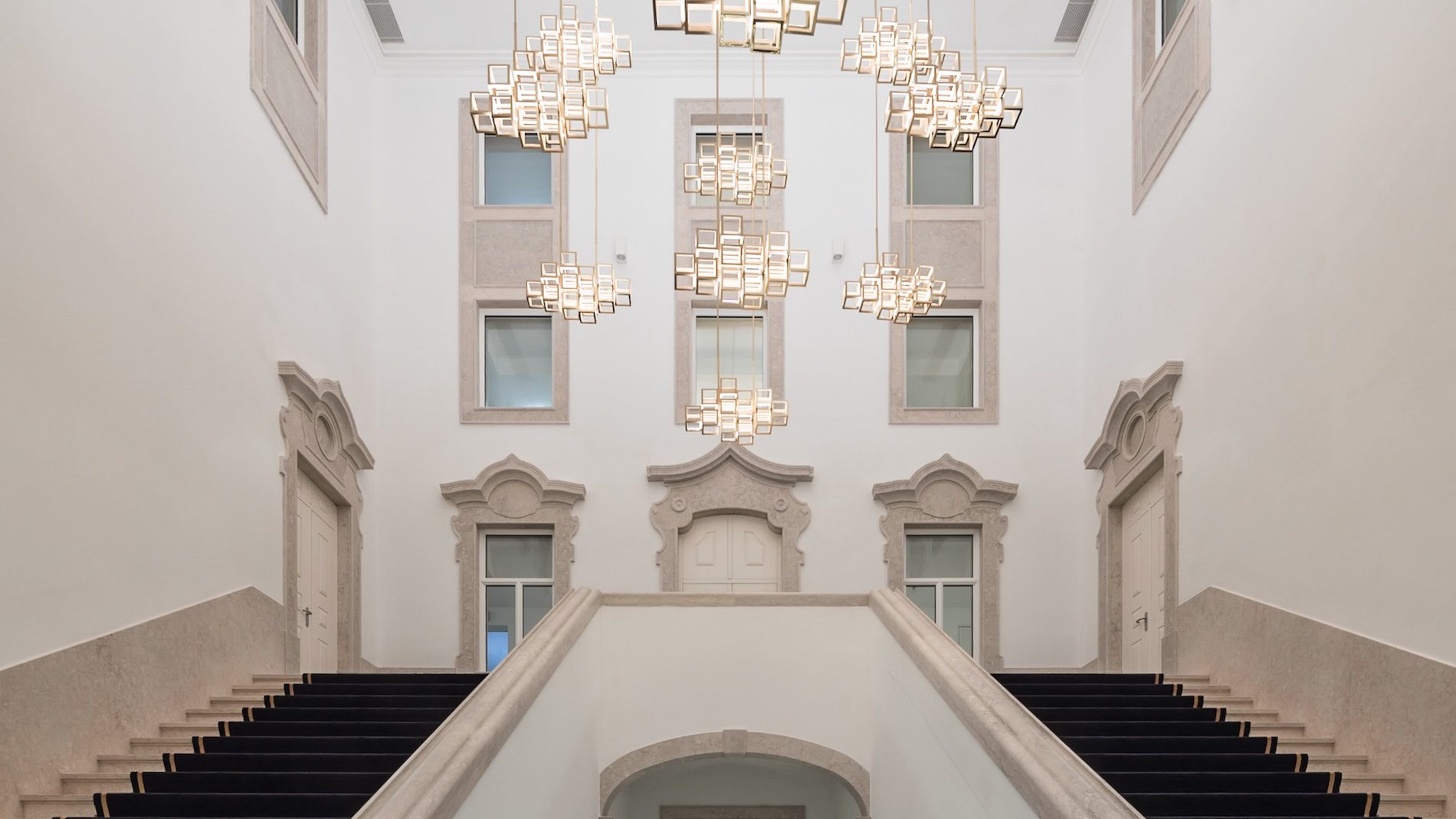 Grand staircase splits into two directions, leading up to ornate doorways and windows. Modern square chandeliers hang from the high ceiling in a bright, white, symmetrical interior with elegant stone details.