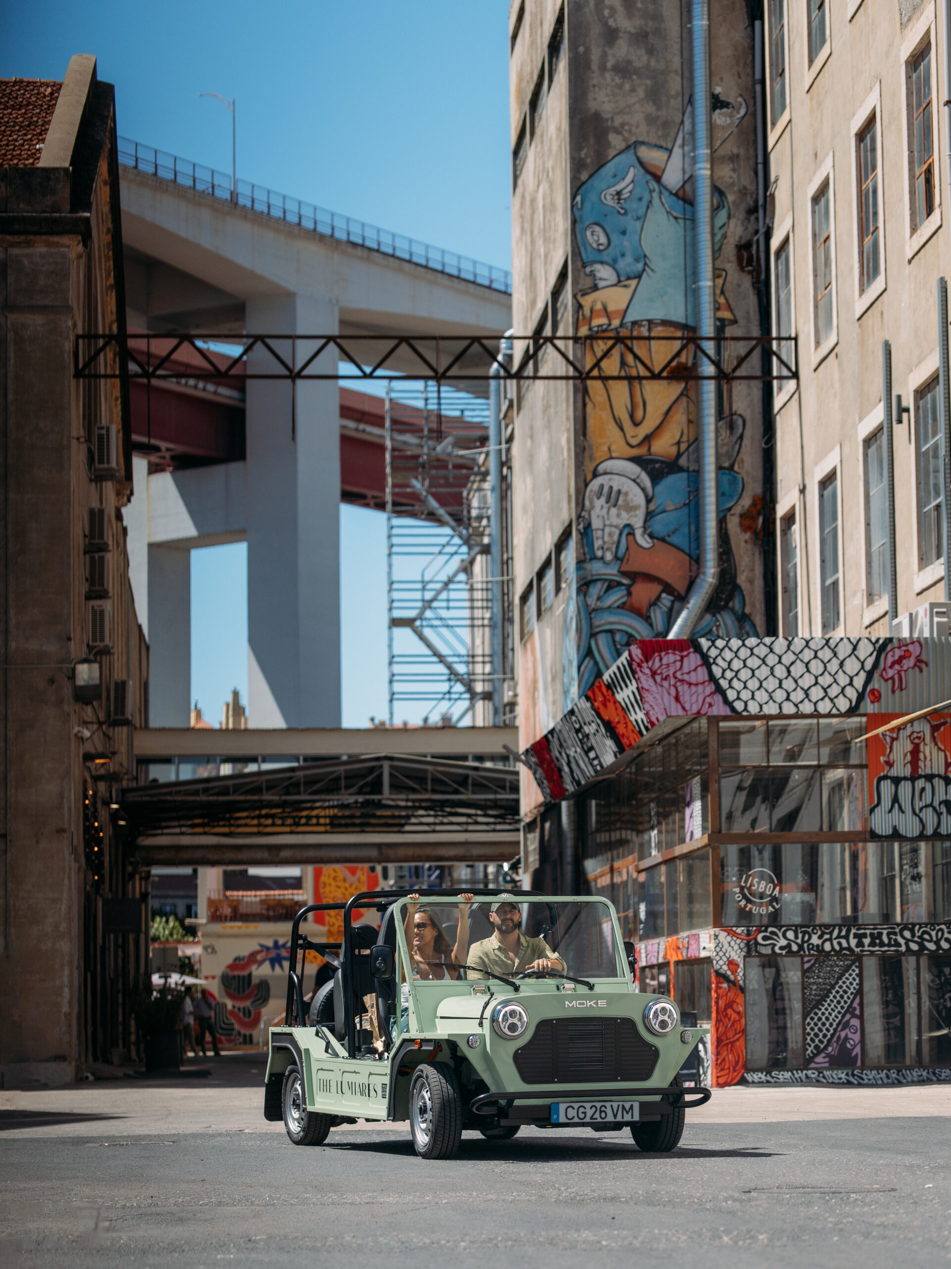 A green open-top jeep with three people inside drives through an urban street lined with tall buildings covered in colorful graffiti. An overpass bridge and scaffolding are visible in the background under a clear blue sky.
