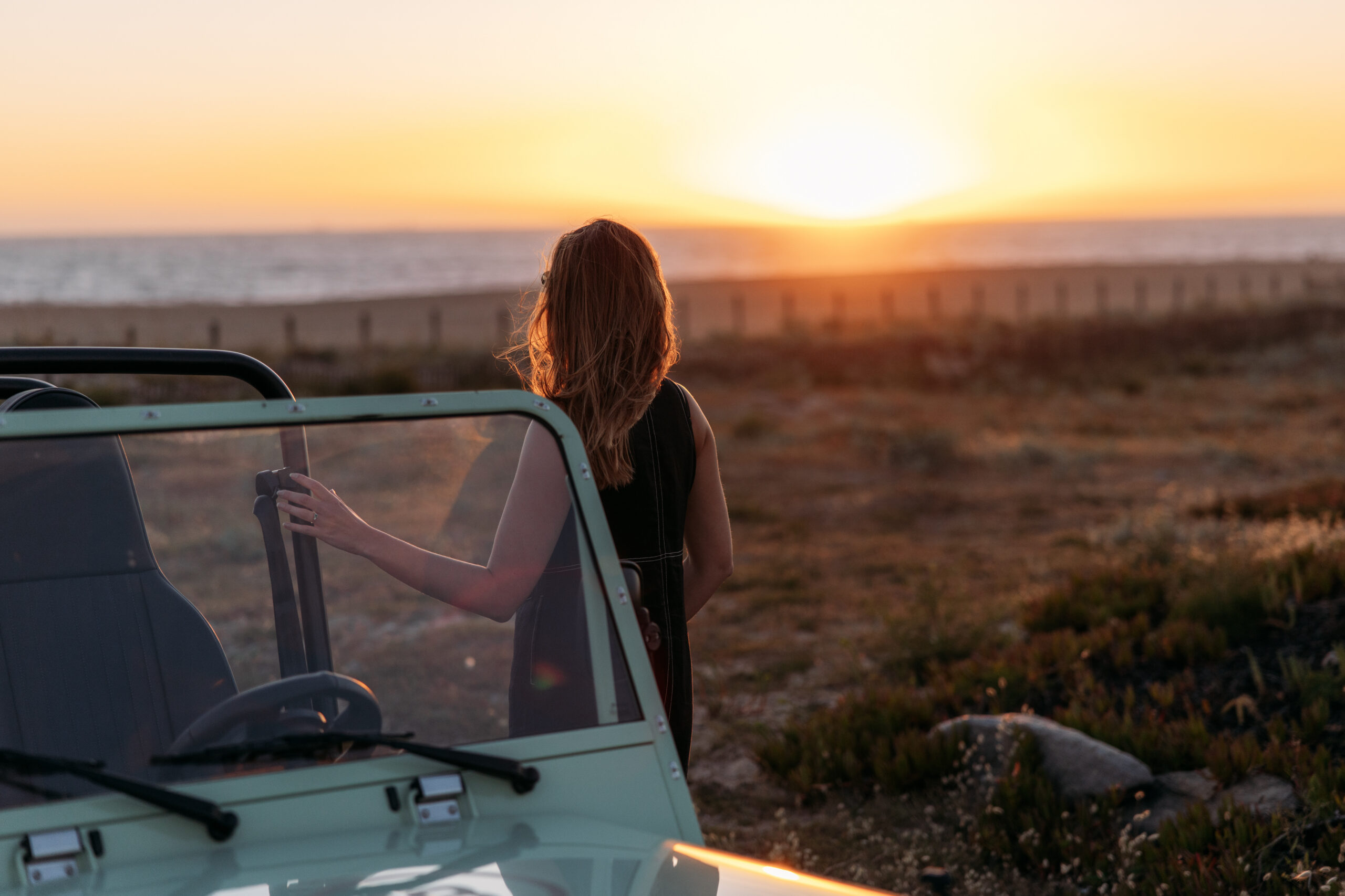 A person with long hair stands next to an open-top vehicle, looking toward the ocean as the sun sets, with warm light illuminating the landscape and sky.